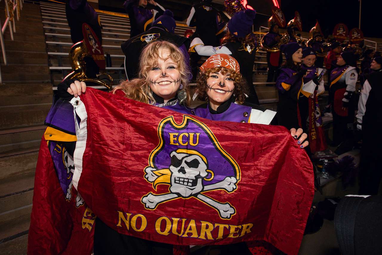 2 members of the ECU Marching Pirates hold a No Quarter flag and smile.